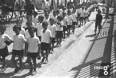 Bambini ed eeducatrici che camminano per una via cittadina.
[La foto  stata scattata a Salerno nel 1937-1943 in via Porto. I bambini in corteo sono diretti verso l'odierna via Ligea allo Stabilimento balneare della colonia marina Mussolini. Una curiosit di questa foto , a destra, sul marciapiede, l'ombra della recinsione che separava il molo 3 gennaio dal porto. Dal profilo dell'ombra si deduce che la recinzione era la stessa bella recinzione metallica, che ancora oggi si pu ammirare e che fu realizzata nel ventennio fascista come si vede dal fascio littorio sagomato sulla superficie alla base di ogni pilastrino circolare e appuntito. Nota di Massimo della Rocca].