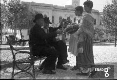 Bambine in abiti tradizionali che offrono prodotti alimentari a persone anziane sedute su una panchina in piazza F.Spirito in occasione della Festa dell'uva